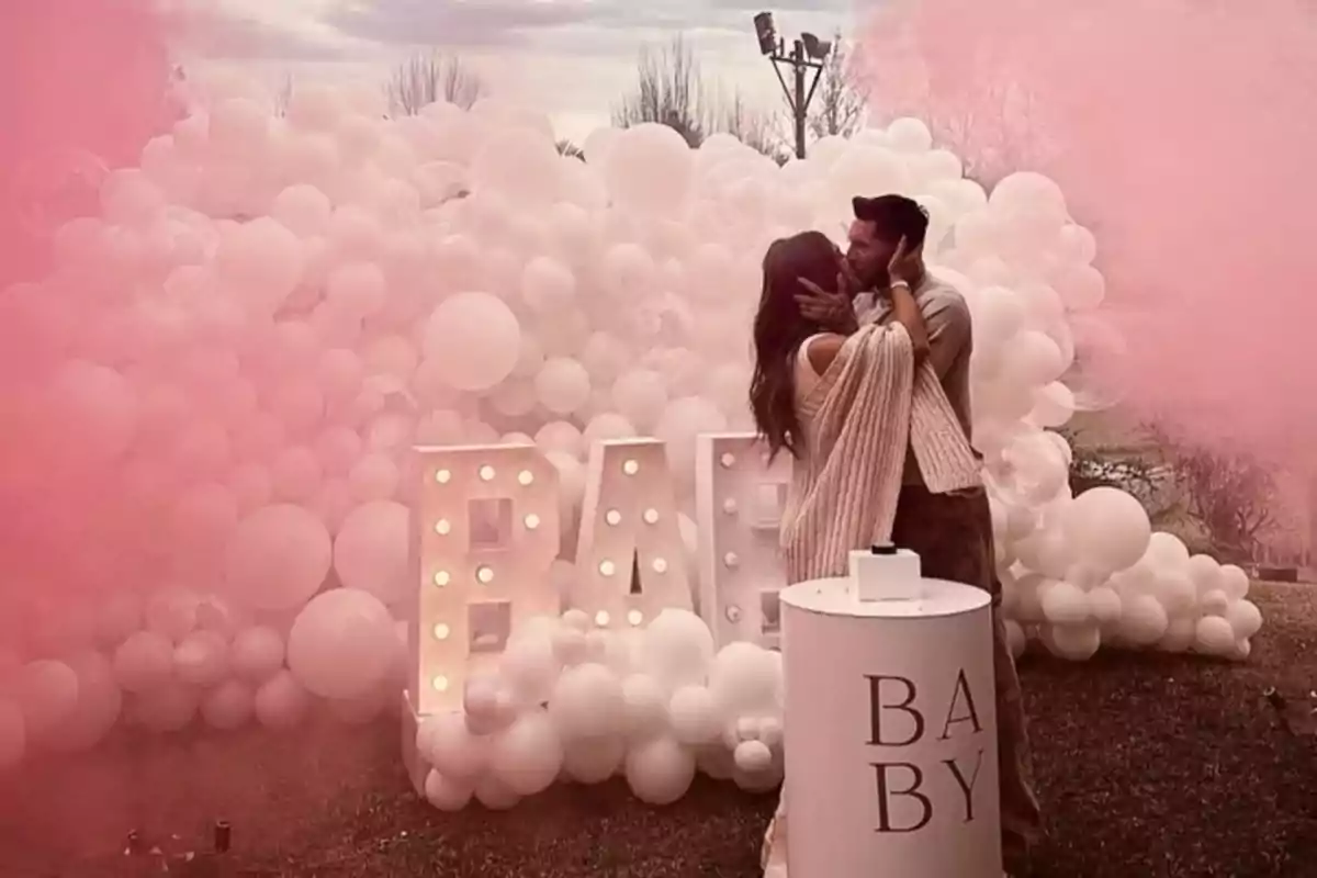 Pareja abrazándose y besándose frente a una decoración de globos blancos y letras iluminadas que forman la palabra BABY con humo rosa a su alrededor Pareja abrazándose y besándose frente a una decoración de globos blancos y letras iluminadas que forman la palabra BABY con humo rosa a su alrededor