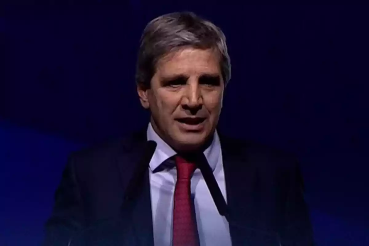 Hombre de cabello canoso con traje oscuro y corbata roja hablando frente a un micrófono sobre un fondo azul oscuro
