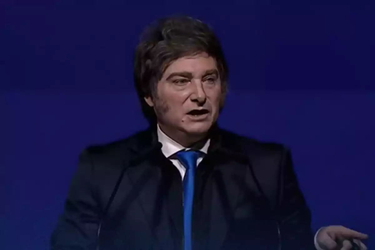 Hombre de cabello castaño con patillas pronunciadas usando traje oscuro y corbata azul hablando frente a un fondo azul