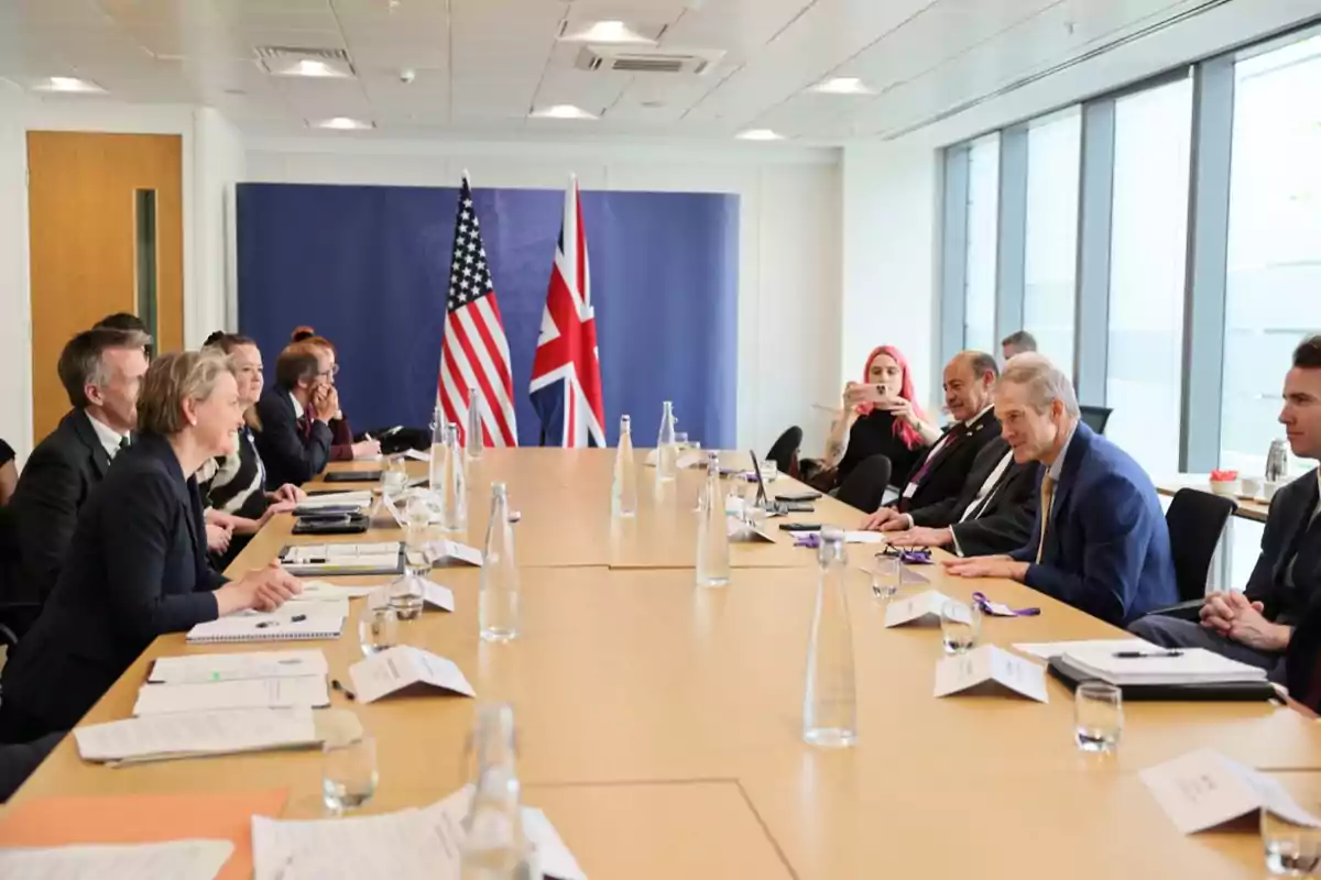 Several people seated around a conference table in a modern room with United States and United Kingdom flags in the background