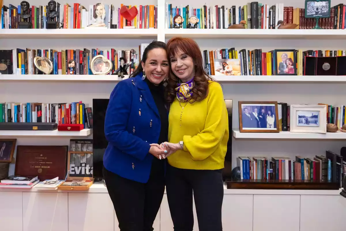 Two women smile and hold hands in front of a shelf filled with books and family photos. Two women smile and hold hands in front of a shelf filled with books and family photos.