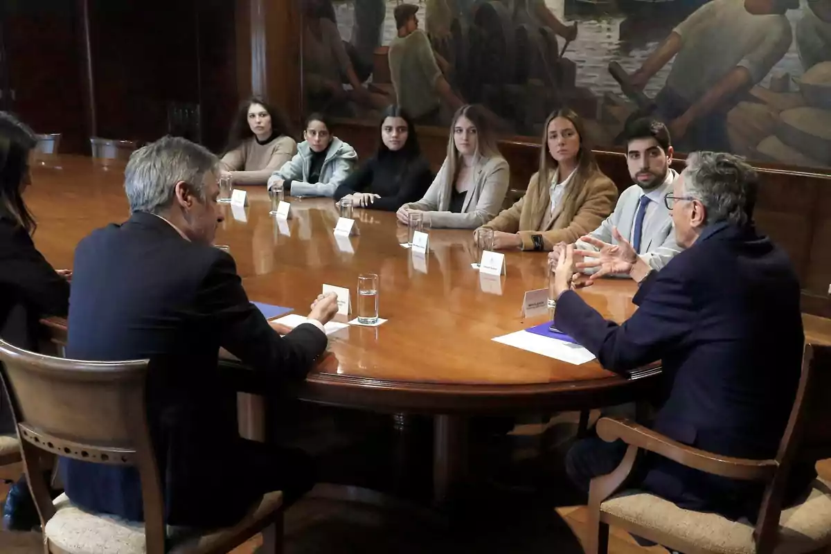 Varias personas sentadas alrededor de una mesa ovalada de madera en una sala de reuniones mientras conversan Varias personas sentadas alrededor de una mesa ovalada de madera en una sala de reuniones mientras conversan
