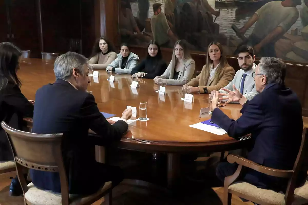 Several people sitting around an oval wooden table in a meeting room while they talk. Several people sitting around an oval wooden table in a meeting room while they talk.