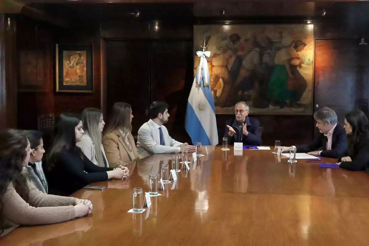 Varias personas sentadas alrededor de una mesa de madera en una sala de reuniones con una bandera de Argentina y cuadros en la pared mientras un hombre mayor habla gesticulando con las manos