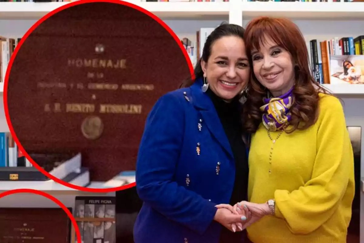 Two women smile and pose with their arms around each other in a library, with a commemorative plaque visible on a bookshelf in the background.