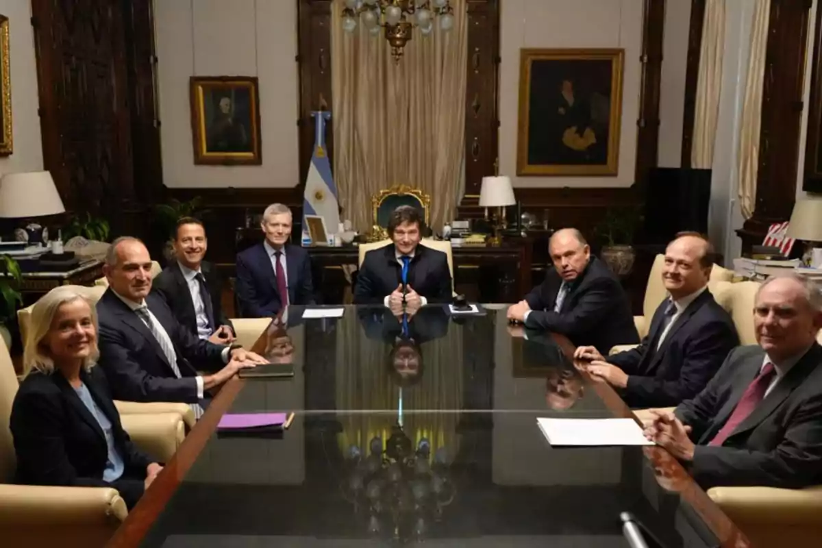 A group of people dressed in suits sitting around a glass table in an elegant room with paintings and an Argentine flag in the background.