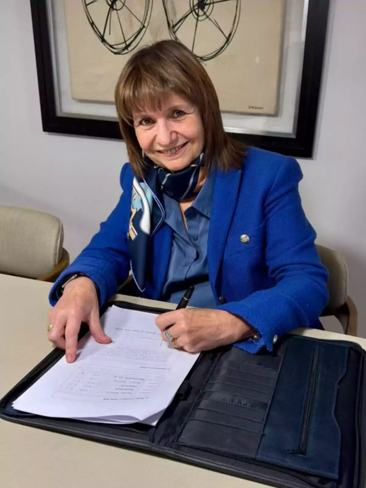 Mujer sonriente con saco azul y pañuelo, sentada en una mesa mientras firma un documento y señala una hoja Mujer sonriente con saco azul y pañuelo, sentada en una mesa mientras firma un documento y señala una hoja