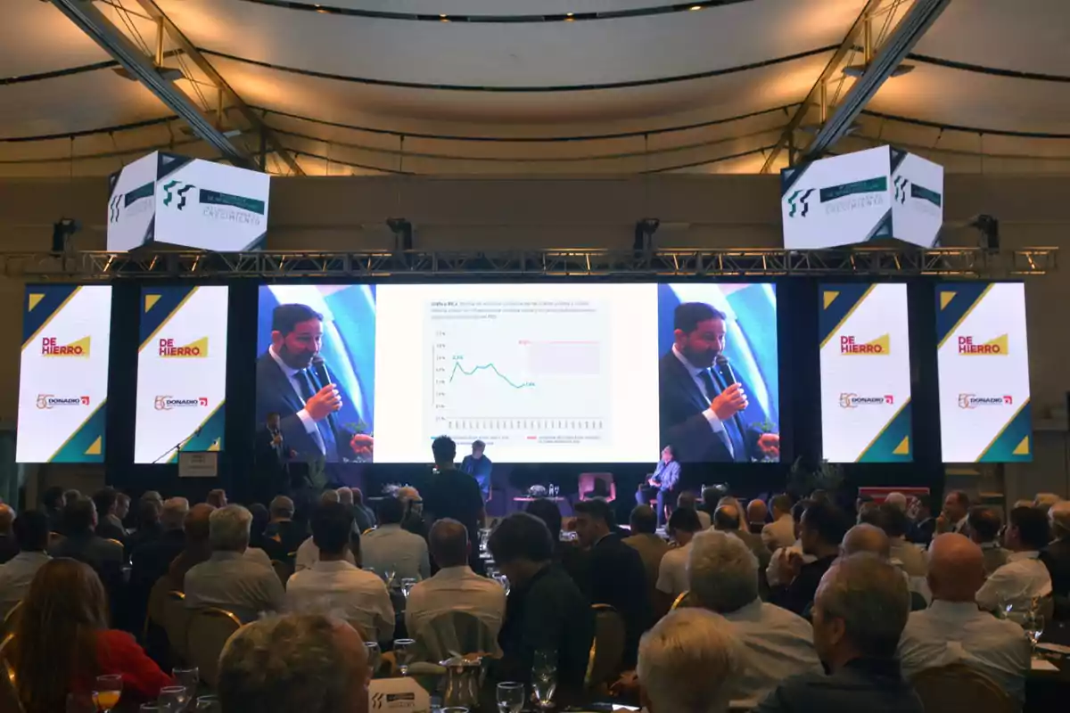An auditorium full of people watches a presentation on a stage with several large screens displaying charts and a speaker talking with a microphone.