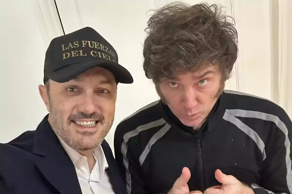 Two men pose for a selfie, one of them wearing a black cap with gold letters and the other giving a thumbs-up gesture.