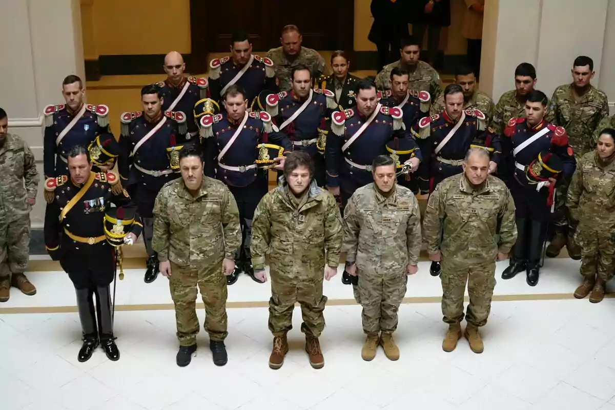 Un grupo de militares y oficiales de diferentes rangos y uniformes están formados en filas dentro de un edificio, algunos con uniformes de gala y otros con ropa de camuflaje, todos mirando hacia el frente en actitud solemne. Un grupo de militares y oficiales de diferentes rangos y uniformes están formados en filas dentro de un edificio, algunos con uniformes de gala y otros con ropa de camuflaje, todos mirando hacia el frente en actitud solemne.