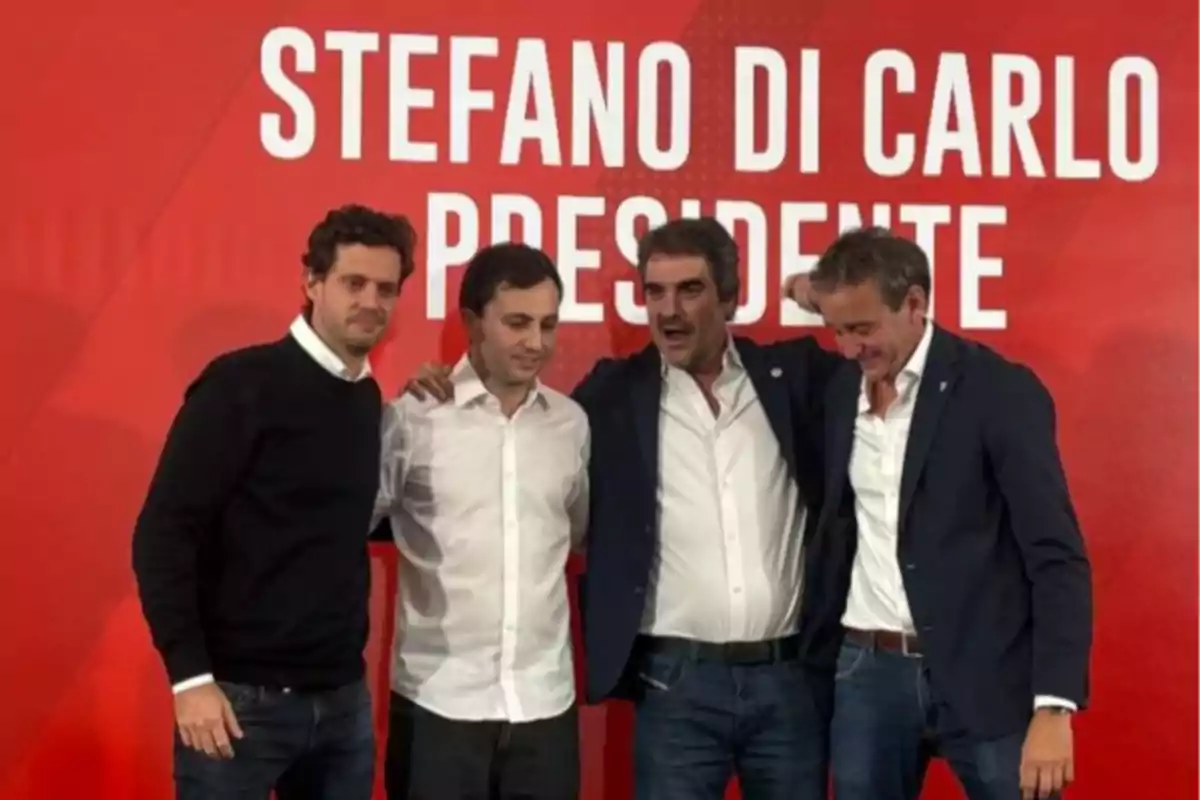 Four men pose together in front of a red background with white letters that say Stefano Di Carlo Presidente. Four men pose together in front of a red background with white letters that say Stefano Di Carlo Presidente.