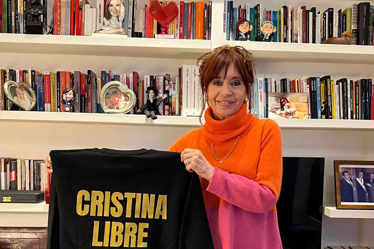 Smiling woman in an orange and pink sweater holds a black garment with gold letters that say CRISTINA LIBRE, with shelves full of books and family photos in the background.