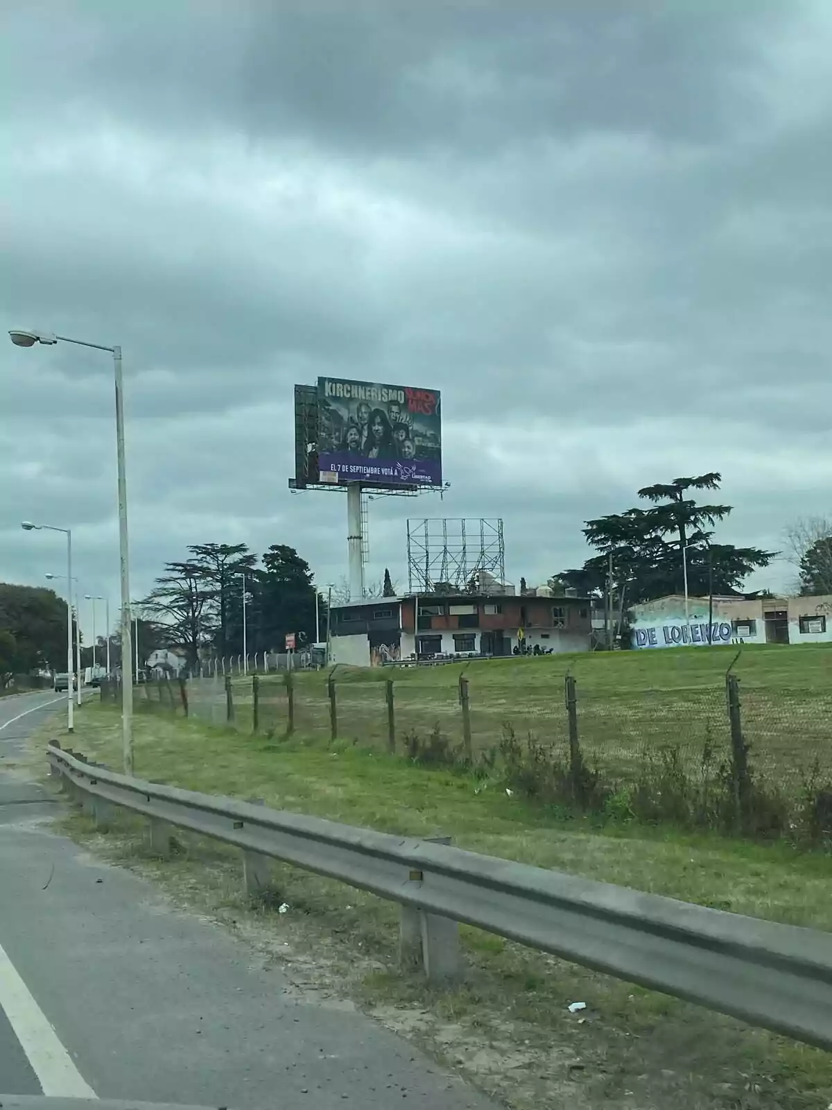 Cartel publicitario grande junto a una carretera en un día nublado con casas y árboles al fondo Cartel publicitario grande junto a una carretera en un día nublado con casas y árboles al fondo