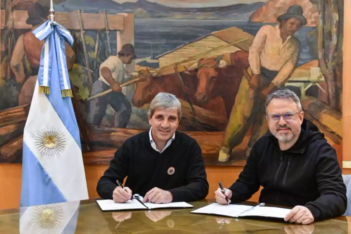 Two men sitting at a table signing documents, with an Argentine flag on the left and a mural in the background depicting rural workers. Two men sitting at a table signing documents, with an Argentine flag on the left and a mural in the background depicting rural workers.