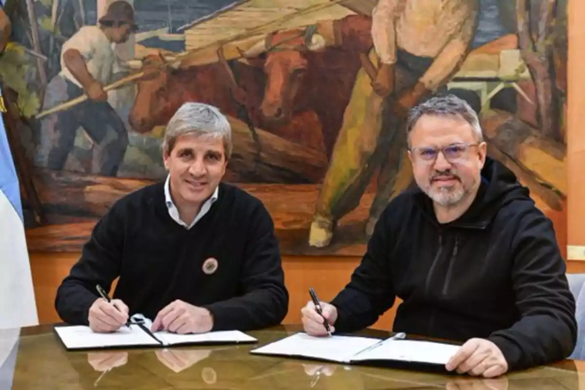 Two men sitting at a table signing documents with a painting in the background depicting a rural scene.