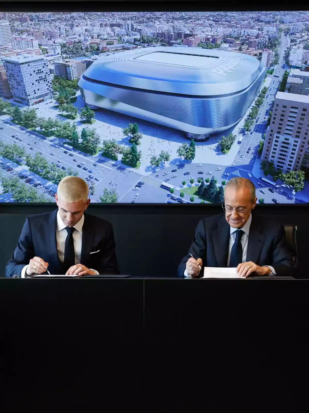 Two men in suits signing documents at a table, with a digital image of a modern stadium projected in the background Two men in suits signing documents at a table, with a digital image of a modern stadium projected in the background