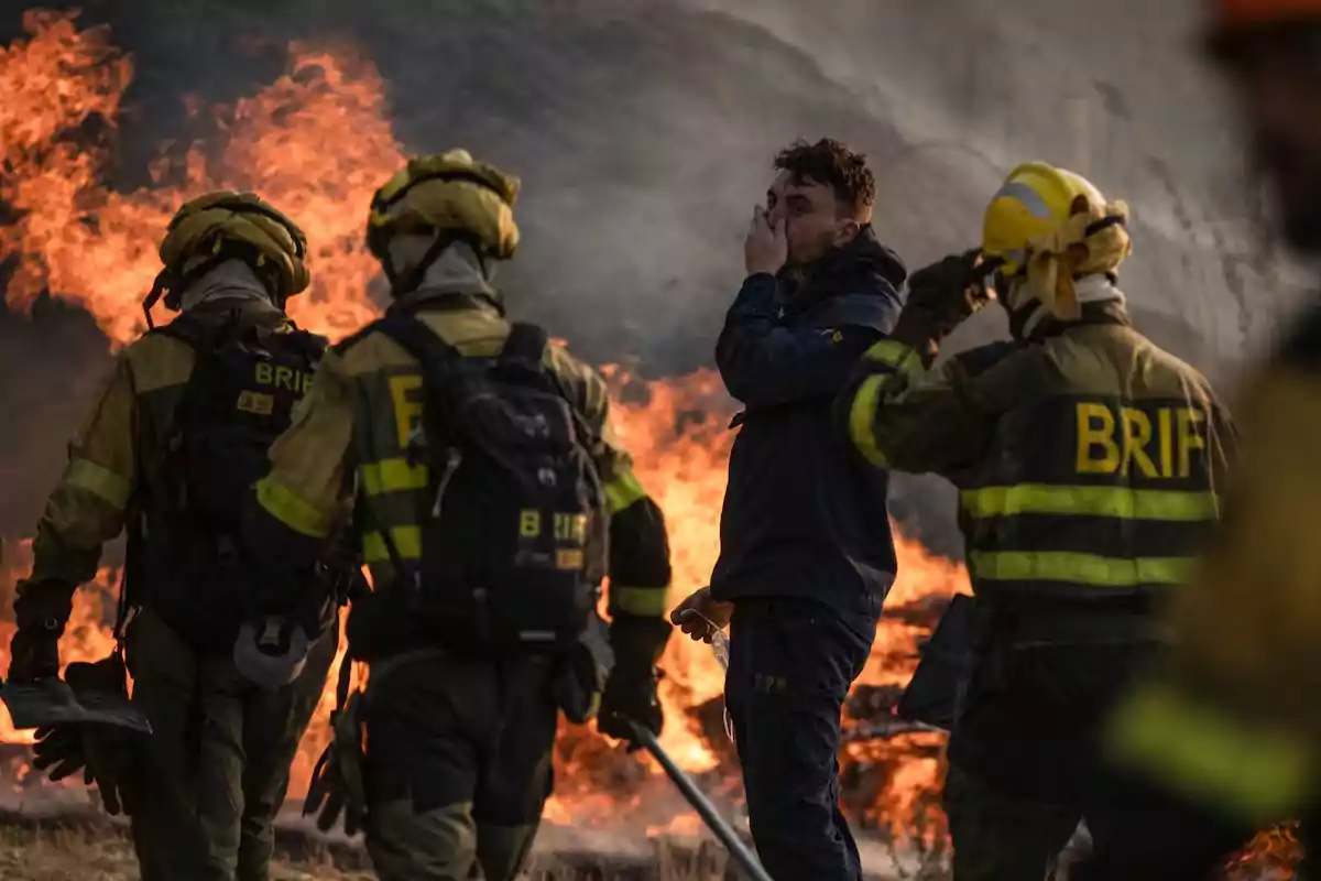 Varios bomberos y un hombre civil se encuentran frente a un incendio forestal mientras el humo y las llamas llenan el fondo