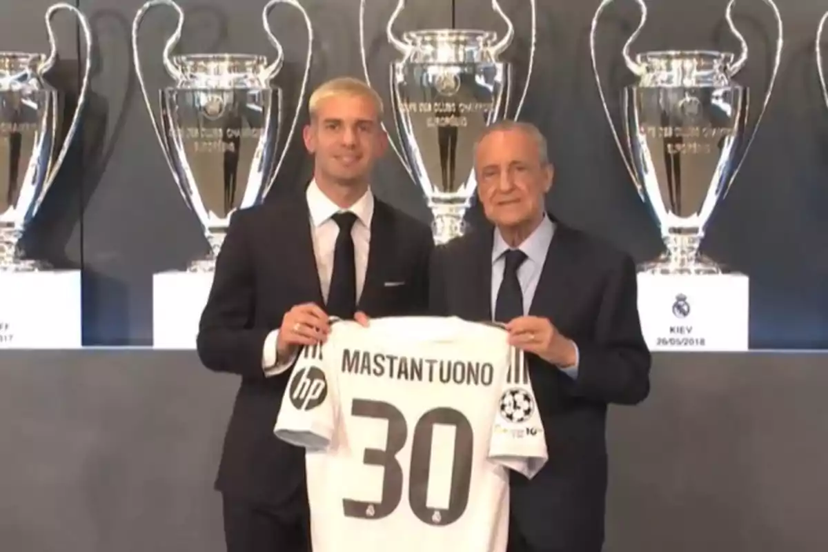 Two men in suits pose holding a soccer jersey with the name Mastantuono and the number 30 in front of several large Champions League trophies.