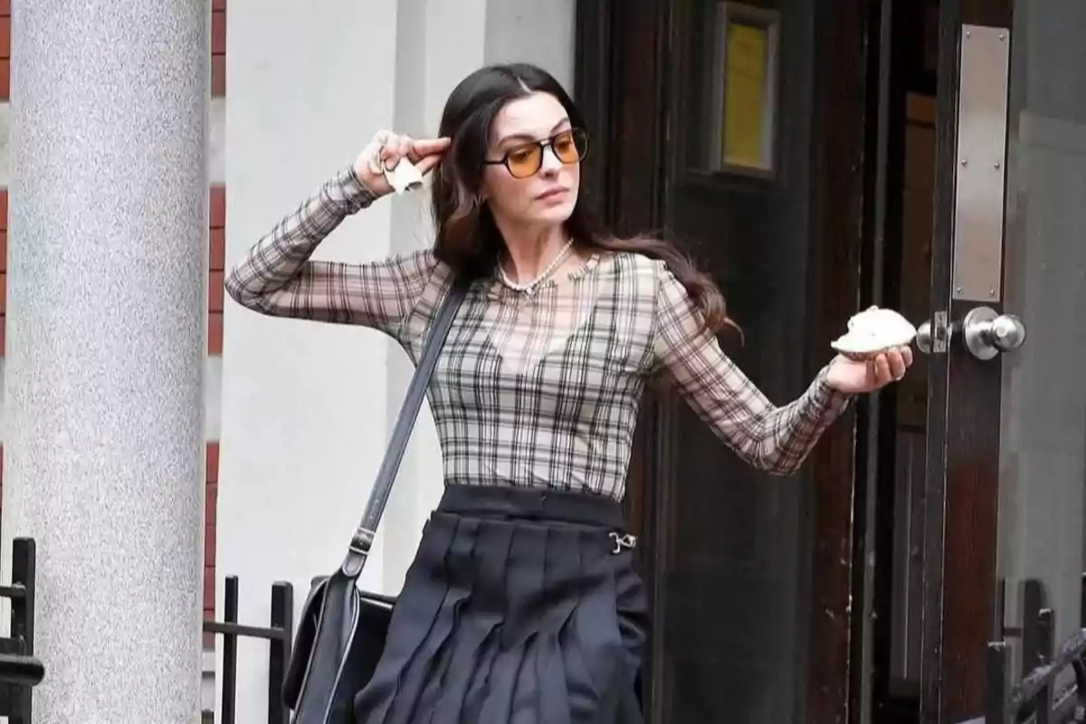 Woman wearing sunglasses and a plaid blouse leaving a building while holding a bun in one hand and touching her head with the other