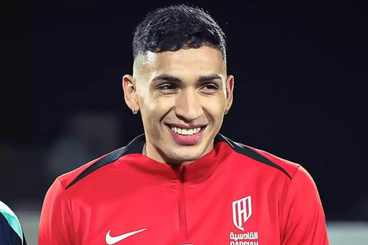 Joven con chaqueta deportiva roja sonríe mientras mira hacia un lado en un entorno oscuro Joven con chaqueta deportiva roja sonríe mientras mira hacia un lado en un entorno oscuro