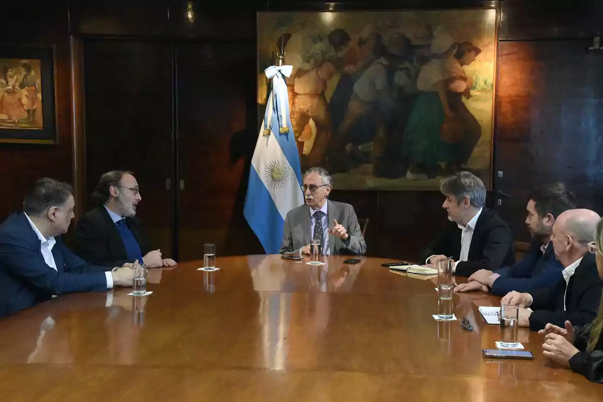 Un grupo de personas en una reunión formal sentadas alrededor de una mesa de madera con la bandera de Argentina y una pintura en la pared de fondo