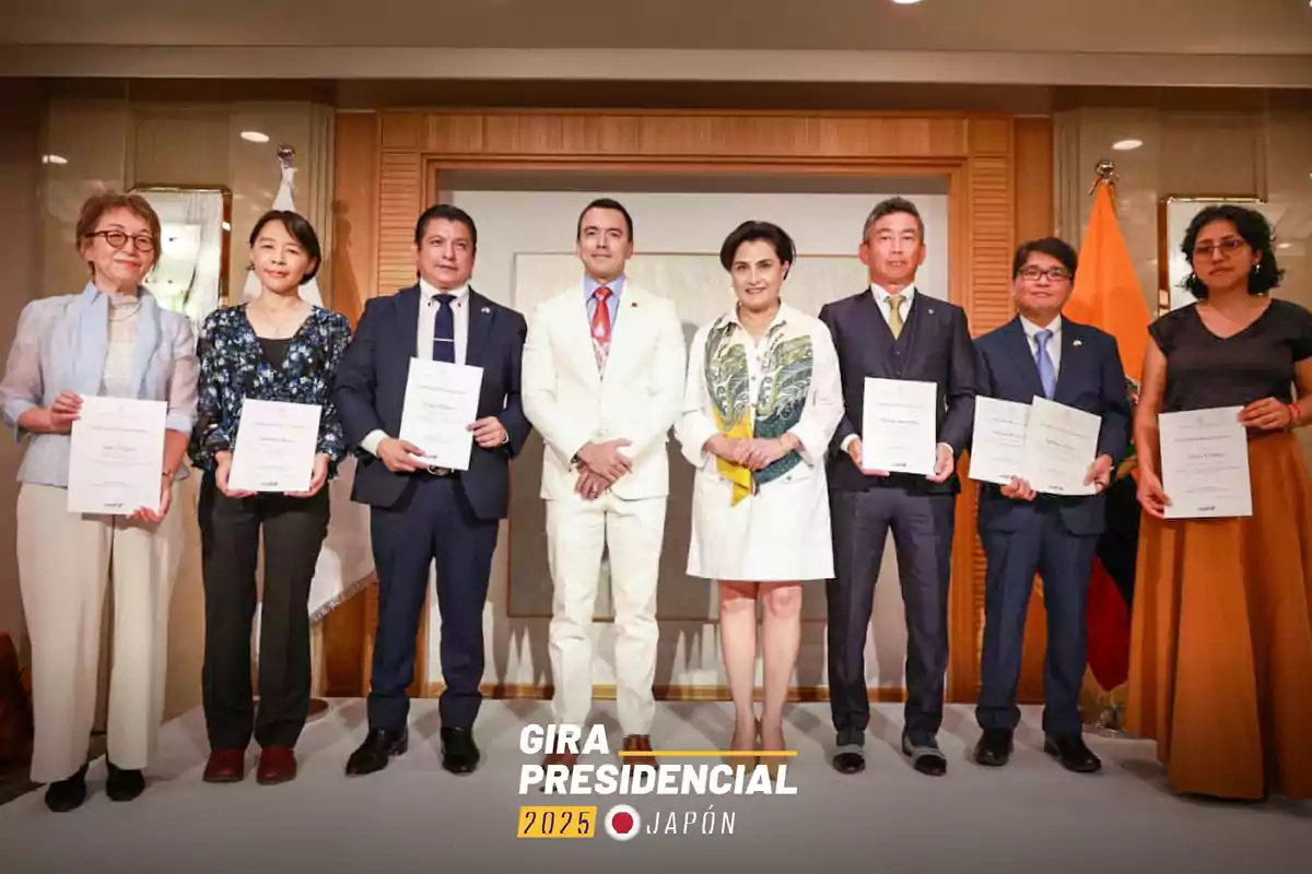 Eight people stand posing in an elegant hall, several of them holding diplomas, and in the background there is an Ecuadorian flag next to a sign that reads Presidential Tour 2025 Japan.