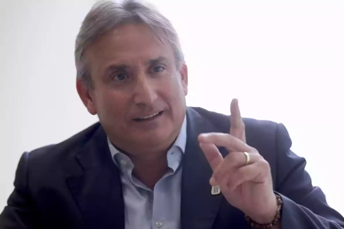 Hombre de cabello canoso con saco oscuro y camisa azul claro levantando el dedo índice mientras habla Hombre de cabello canoso con saco oscuro y camisa azul claro levantando el dedo índice mientras habla