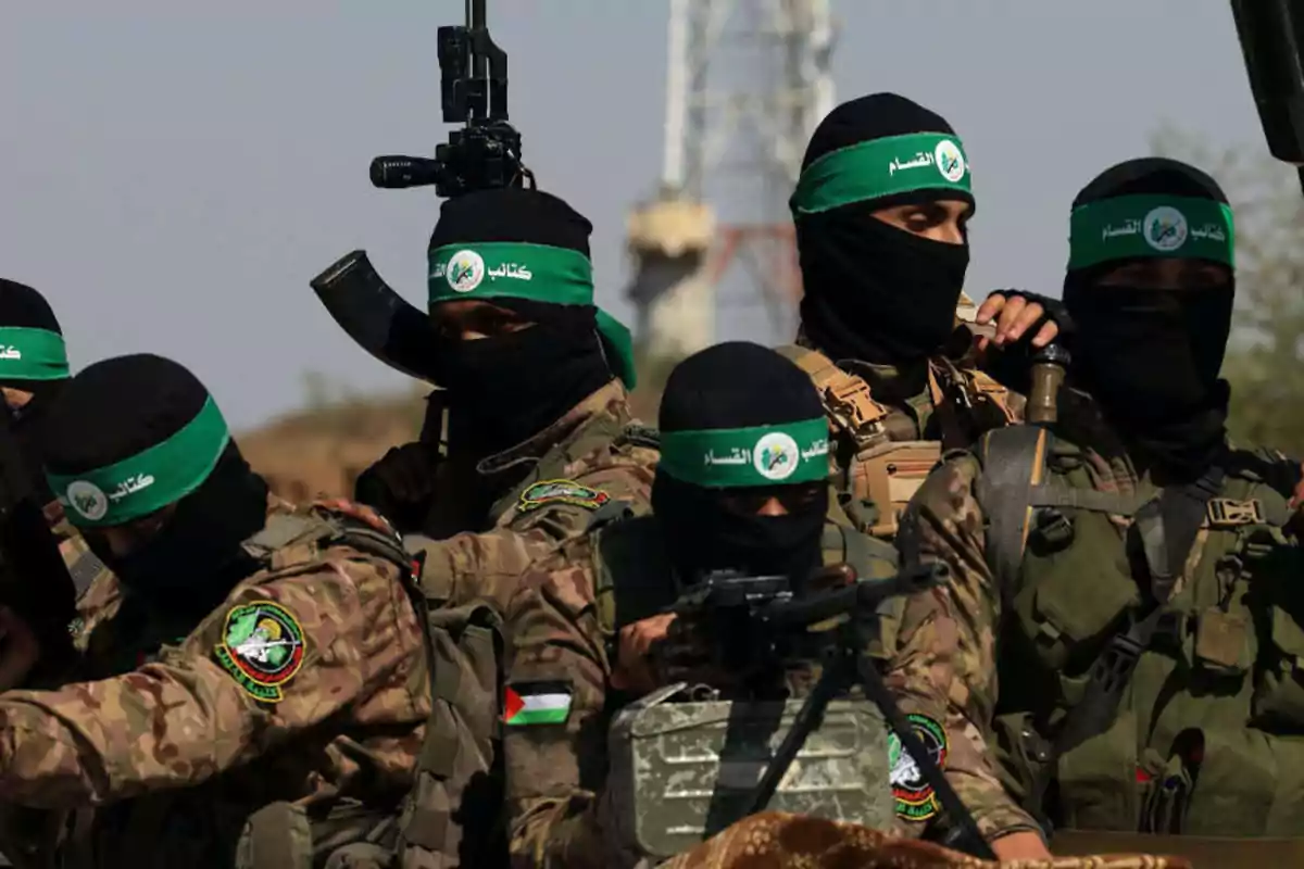 Varios hombres armados con uniformes de camuflaje y pasamontañas negros con cintas verdes en la cabeza se agrupan en un entorno exterior