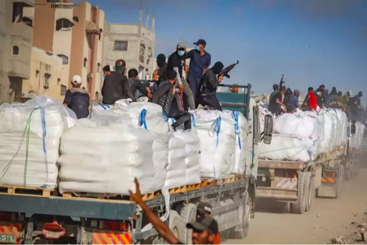 Trucks loaded with white sacks move along a dusty street while several armed, masked people ride on top of the cargo in an urban area.