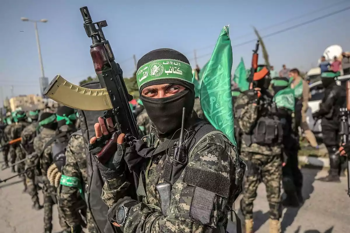 Un grupo de hombres armados con uniformes de camuflaje y pañuelos verdes en la cabeza se encuentra reunido al aire libre, uno de ellos mira directamente a la cámara sosteniendo un rifle.