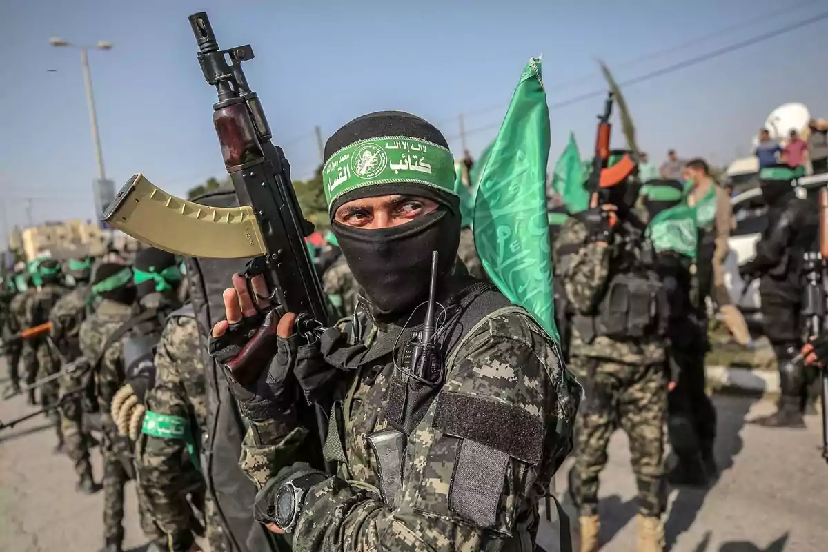 A group of people dressed in camouflage uniforms and green scarves hold weapons and green flags on a street during a demonstration or parade.