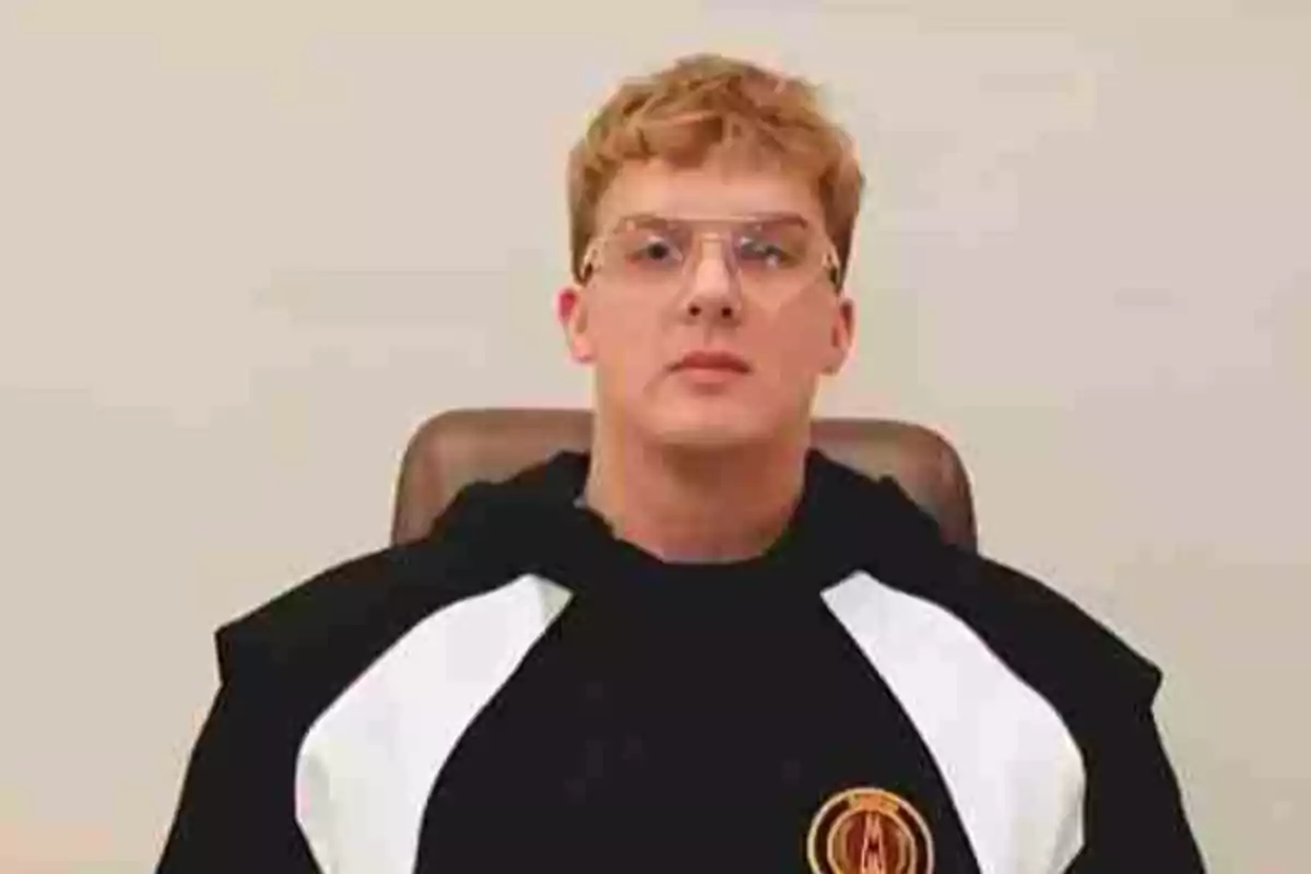 Young man with short blond hair and clear glasses sitting on a chair, wearing a black and white sweatjersey with a circular logo on the chest Young man with short blond hair and clear glasses sitting on a chair, wearing a black and white sweatjersey with a circular logo on the chest