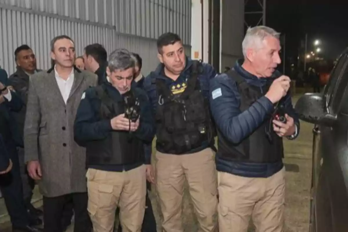 Grupo de hombres con chalecos antibalas y radios de comunicación junto a un vehículo en un entorno industrial por la noche Grupo de hombres con chalecos antibalas y radios de comunicación junto a un vehículo en un entorno industrial por la noche