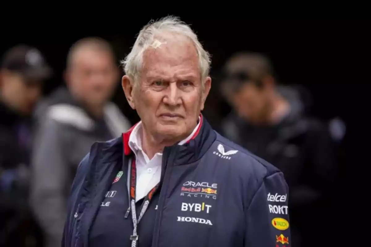 Older man wearing a Red Bull Racing jacket with a serious expression in an outdoor setting with blurred people in the background Older man wearing a Red Bull Racing jacket with a serious expression in an outdoor setting with blurred people in the background