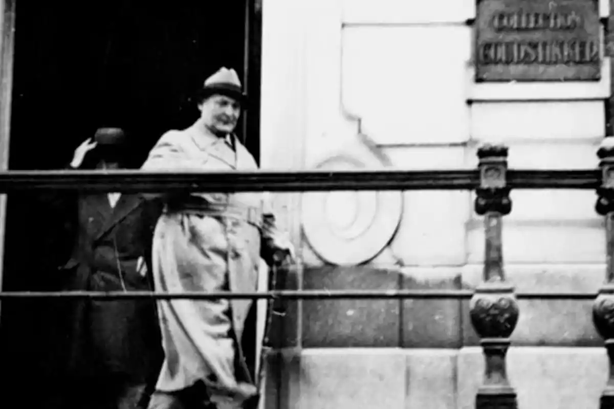 Two people wearing coats and hats are coming out of an old building, one of them is in the foreground and the other in the background; the image is in black and white, and a railing and a plaque on the wall can be seen. Two people wearing coats and hats are coming out of an old building, one of them is in the foreground and the other in the background; the image is in black and white, and a railing and a plaque on the wall can be seen.