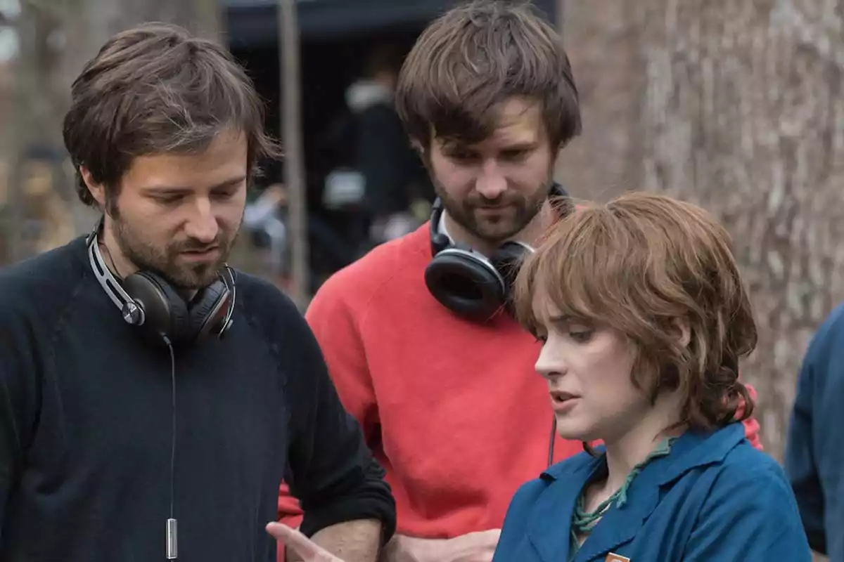 Three people are having a focused conversation on a film set surrounded by trees; two of them have headphones around their necks and one is wearing blue overalls. Three people are having a focused conversation on a film set surrounded by trees; two of them have headphones around their necks and one is wearing blue overalls.