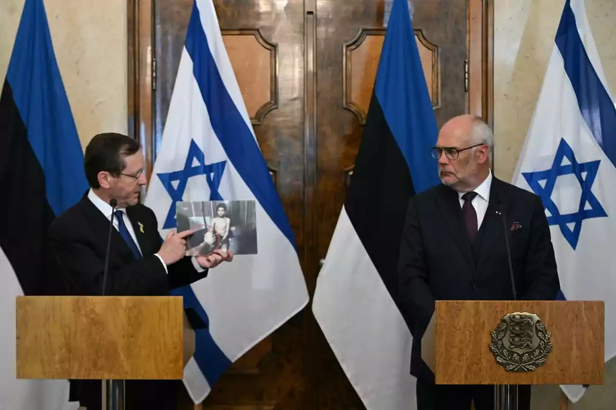 Dos hombres de traje están de pie en atriles frente a banderas de Estonia e Israel uno de ellos sostiene y muestra una fotografía mientras el otro lo observa