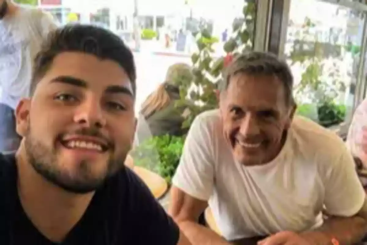 Dos hombres sonríen mientras se toman una selfie en lo que parece ser una cafetería iluminada por la luz natural que entra por las ventanas. Dos hombres sonríen mientras se toman una selfie en lo que parece ser una cafetería iluminada por la luz natural que entra por las ventanas.