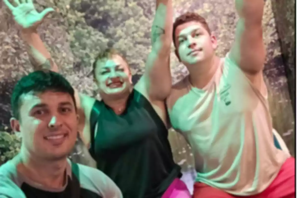 Tres personas sonríen y posan juntas frente a un fondo de vegetación, dos de ellas levantan los brazos mientras la persona a la izquierda mira a la cámara