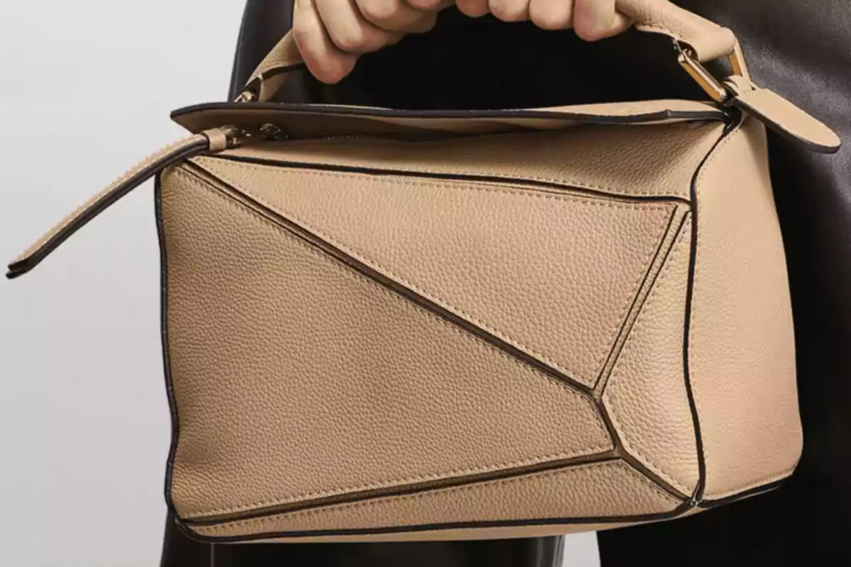 Bolso de cuero beige con diseño geométrico y asa corta sostenido por una mano Bolso de cuero beige con diseño geométrico y asa corta sostenido por una mano