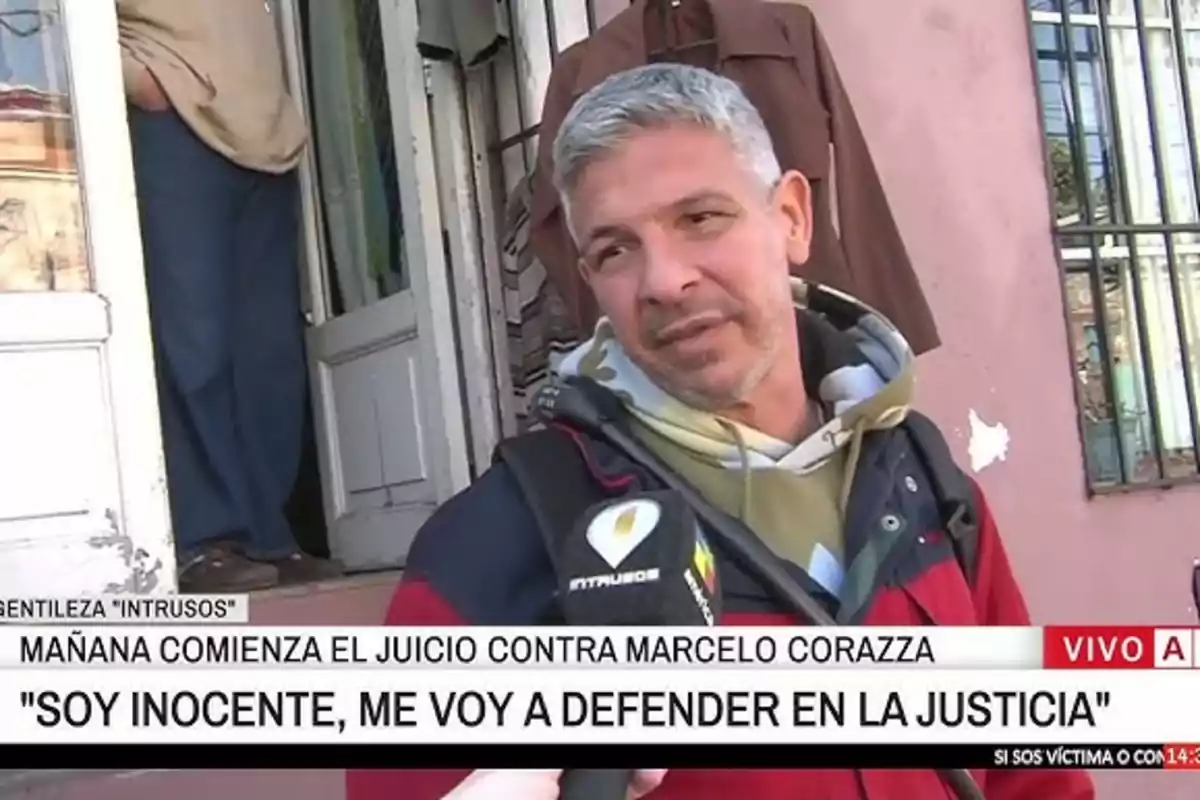 Gray-haired man with a short beard being interviewed by a television channel in front of a house with an open door and a window on the pink wall, with on-screen text mentioning the start of a trial and a declaration of innocence.