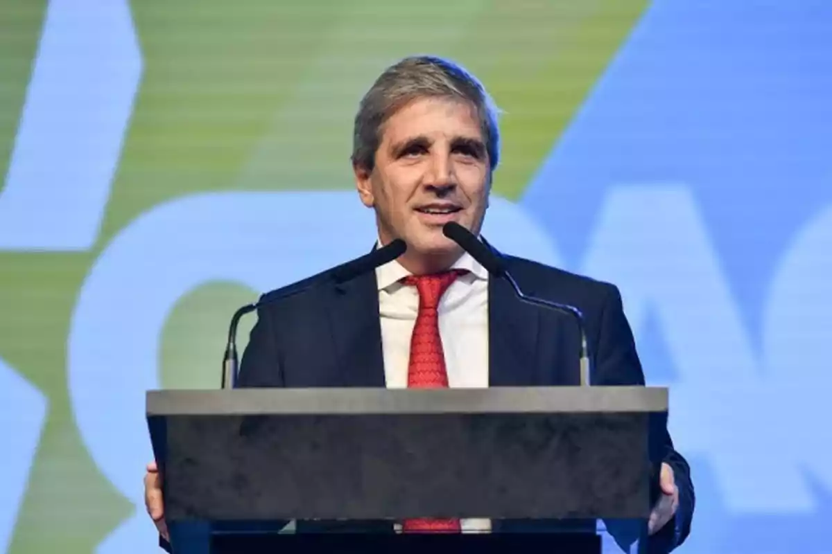 Hombre de cabello canoso con traje oscuro y corbata roja hablando en un podio con dos micrófonos frente a un fondo azul y verde Hombre de cabello canoso con traje oscuro y corbata roja hablando en un podio con dos micrófonos frente a un fondo azul y verde