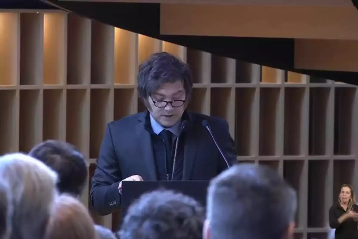 Man with glasses and a dark suit speaking at a podium in front of an audience in a modern auditorium Man with glasses and a dark suit speaking at a podium in front of an audience in a modern auditorium