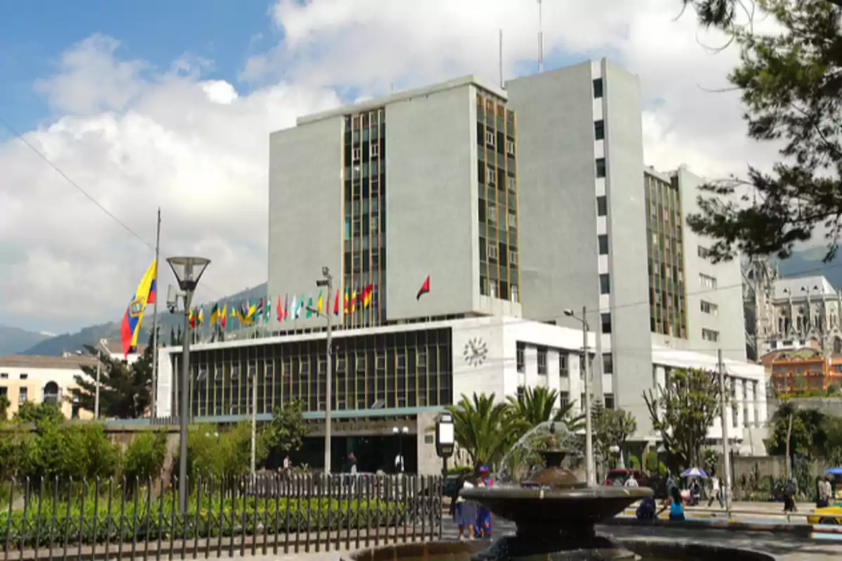 Edificio gubernamental de varios pisos con banderas en la parte superior y una fuente en primer plano rodeada de vegetación