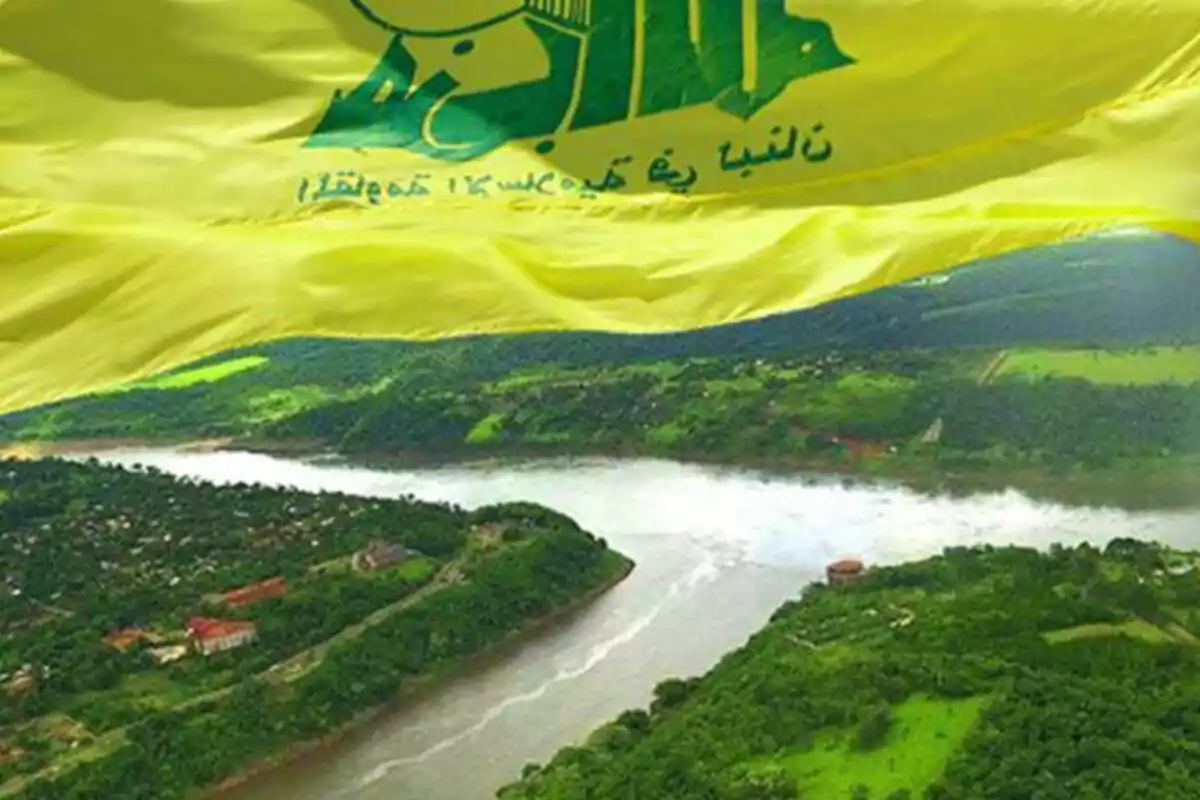 Bandera amarilla con símbolos y letras en árabe ondeando sobre un paisaje de río y vegetación abundante