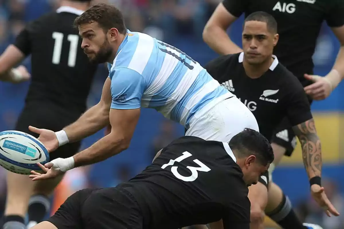 Rugby player in a light blue and white uniform passes the ball while being tackled by an opponent dressed in black during a match.