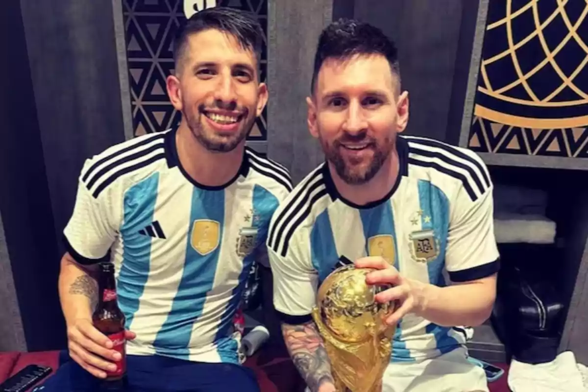 Dos futbolistas argentinos sonríen en un vestuario, uno sostiene una botella y el otro el trofeo de la Copa del Mundo Dos futbolistas argentinos sonríen en un vestuario, uno sostiene una botella y el otro el trofeo de la Copa del Mundo