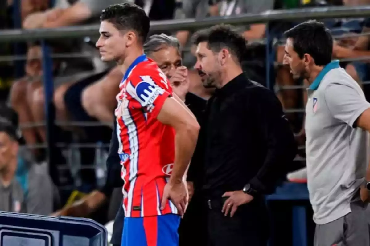 Un jugador de fútbol con uniforme rojo y blanco está en la banda junto a tres personas del cuerpo técnico durante un partido Un jugador de fútbol con uniforme rojo y blanco está en la banda junto a tres personas del cuerpo técnico durante un partido