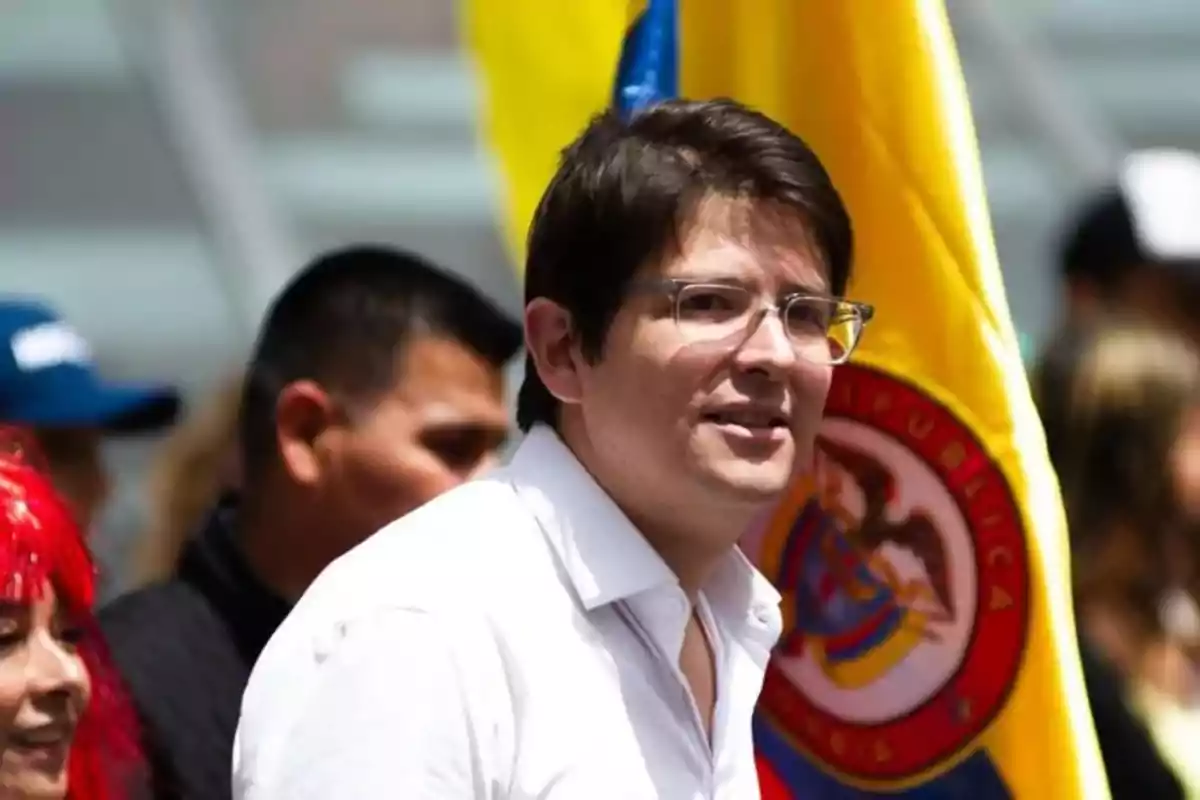 Hombre con gafas y camisa blanca frente a una bandera de Colombia en un evento al aire libre