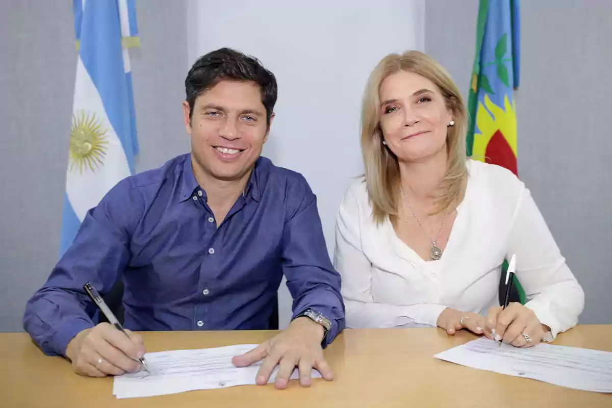Two people smile while signing documents at a table, with the flags of Argentina and the province of Buenos Aires in the background.